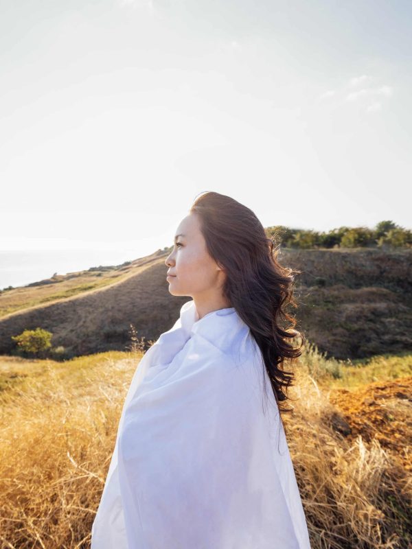 A young handsome Asian woman wrapped in a white sheet and looks at the seascape. A charming Korean woman enjoys nature. Profile of an attractive girl in the rays of the sun at sunset outdoors.