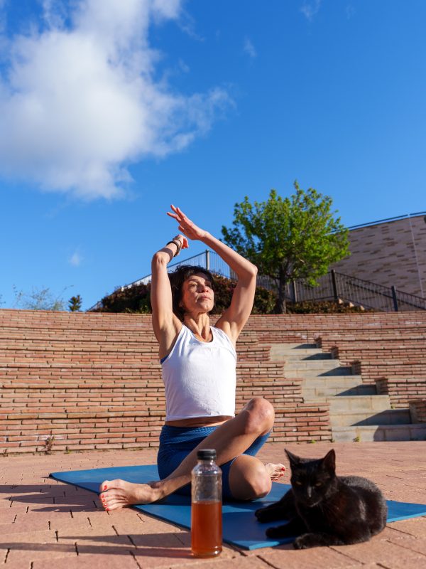 Mature woman practicing yoga in a sunlit park, enjoying a moment of tranquility while her black cat relaxes peacefully on the mat