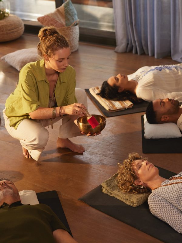 Woman leading sound healing session with singing bowl at indoor wellness retreat. Group of multiethnic people lying down in deep relaxation on yoga mats, surrounded by crystals and warm natural light. Peaceful moment of energy work with people lying on back in circle while the holistic therapist plays tibetan bells.