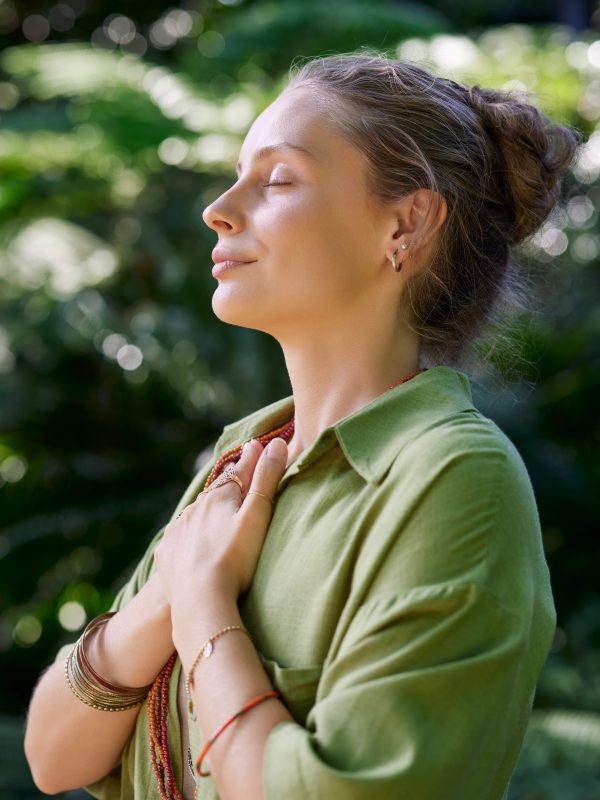 Woman with closed eyes and arms on chest practicing mindfulness while enjoying summer retreat camp. Woman practices meditation and emotional grounding in a peaceful retreat setting. A calm girl gently holds her hands over her heart, embracing gratitude and stillness amidst nature.